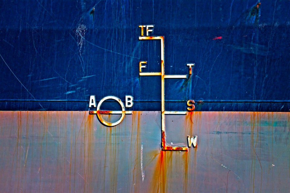 A closeup photo of the side of a container ship, showing a series of lines. On the left is a circle intersected horizontally by a line, the ends of which are labelled A and B. To the right of that is a vertical line with several horizontal lines branching off from it -- the top one is labeled TF, the ones below it are labeled F and T, the one below that is labelled S, and the bottom one is labeled W. The ship is painted blue in the top half of the photo and grey in the bottom half; both colors are corroded slightly by weather, with the grey part adorned with orange streaks of rust