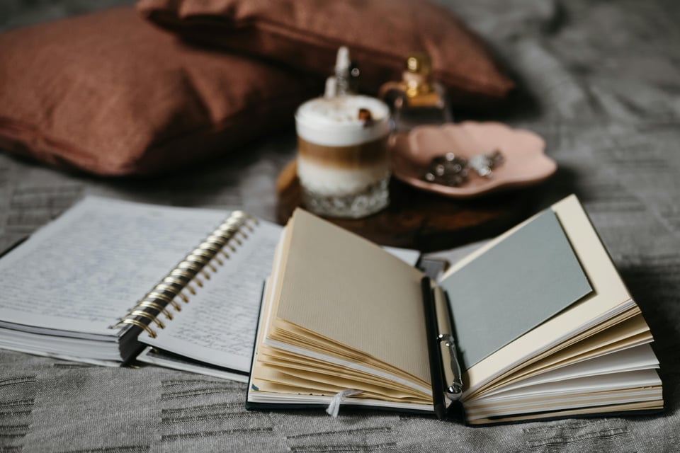 Stock image of a few notebooks lying on a blnket with a pen inbetween one notebook's pages. The background shows some comfy pillows and a small tray with accessories and a small drink on it.