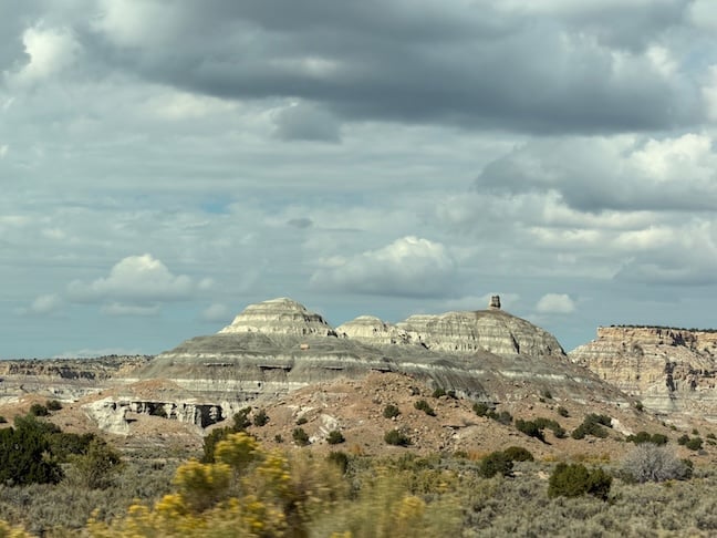 Large mountainous stratified sandstone layers near New Mexico's Badlands, Angel Peak Scenic Area along Route 550 north. Clouds in the sky. Desert yellow flowers bloom.