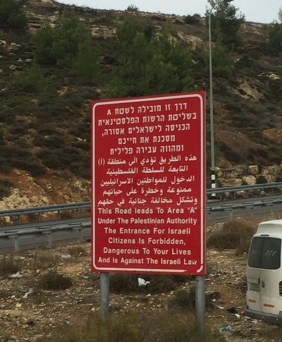 A red sign on the side of a road that says in Hebrew, Arabic and English:  "This Road leads to Area 'A' Under the Palestinian Authority. The Entrance for Israeli Citizens is Forbidden, Dangerous To Your Lives And Is Against The Israeli Law"