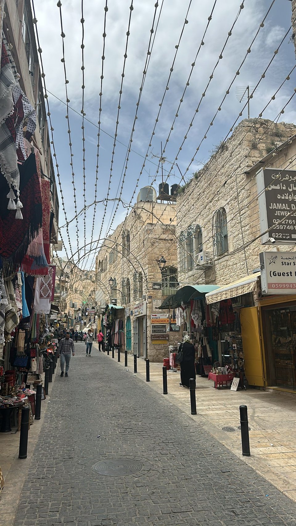 A long cobblestone street with just a few people, shops on the sides of the street. Lights strung atop the walkway.et