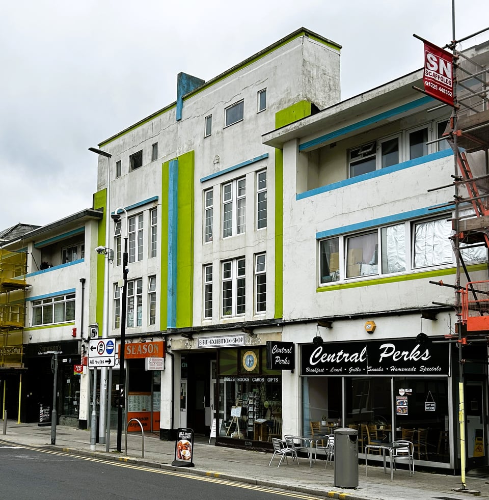 The central feature of a 1930s parade of shops with flats over. The central part is four storeys high and two shops wide. It has neat rows of windows and a central fin detail. To either side, the building drops to being three storeys high, with inset balconies on the top floors. The deco details have been picked out against the white render in an acid green and bright blue. One of the shops is a cafe called Central Perks.