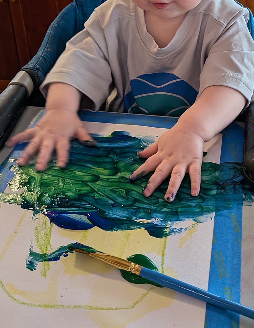 A young toddler is using both hands to smear blue and green paint across a piece of paper taped to the table; there is an abandoned paintbrush trailing paint in the foreground. It's a giant mess but she's having a great time; from the tiny bit of her face that's visible, she's smiling.