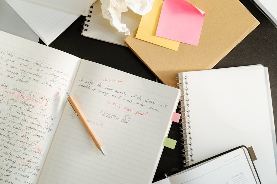 Image shows a pen lying on top of an open notebook, surrounded by several other notebooks and sticky notes.