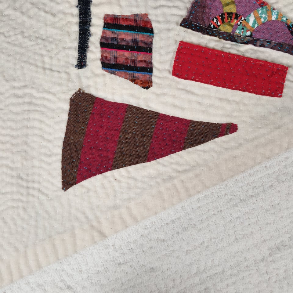 Detail view of Meet Me at the Corner in progress. This view has several pieces of fabric in reds and purples against an off-white background. Sarah's hand stitches are visible.
