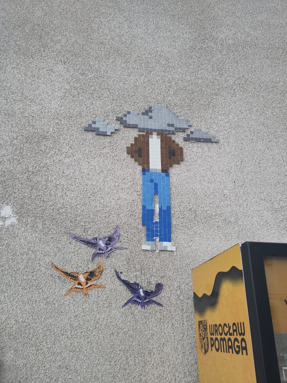Wandbild mit einem menschen, dessen Kopf von Wolken verdeckt ist. Dazu sind drei flatternde Vögel abgebildet. Vor der Wand steht ein Automat, auf dem "Wroclaw Pomaga" gedruckt ist.