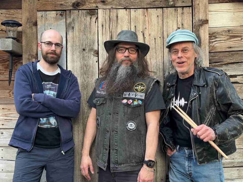 A balding man with a beard and glasses stands next to a man with a prodigious beard in a faded black denim vest, who stands next to a man wearing a beat-up black leather jacket and holding a pair of drums sticks