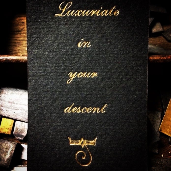 A letterpress card, black with gold cursive that reads: Luxuriate in your descent. A decorative design at the bottom.