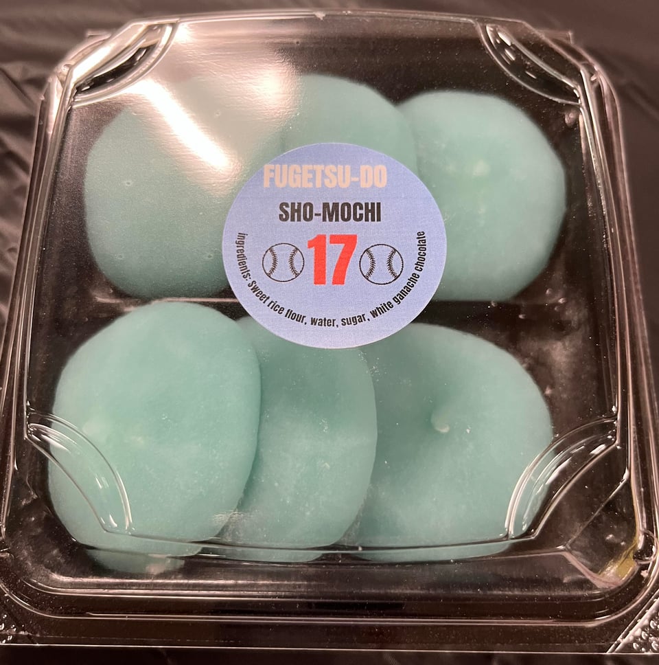 A square black plastic package with a clear top containing six pieces of blue mochi. A round blue sticker in the center of the package reads: Fugetsu-do Sho-Mochi 17.