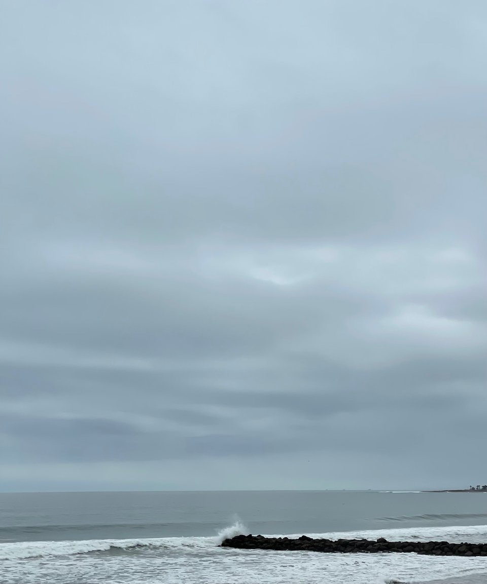 A large overcast sky dominates most of the frame, at the very bottom a wave explodes on the tip of the breaker