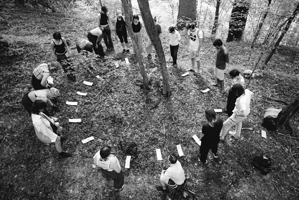 [A black and white photo of people standing in a big circle in the woods. In the center of their circle is a tree, and placed in front of each person’s feet is a long vertical rectangle of paper.]