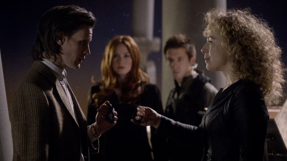 The Doctor gets married to River Song. He's wearing his tweed jacket and holding an object in his hand. River is wearing a leather jacket and holding out her hand. In the background, Rory and Amy are watching.