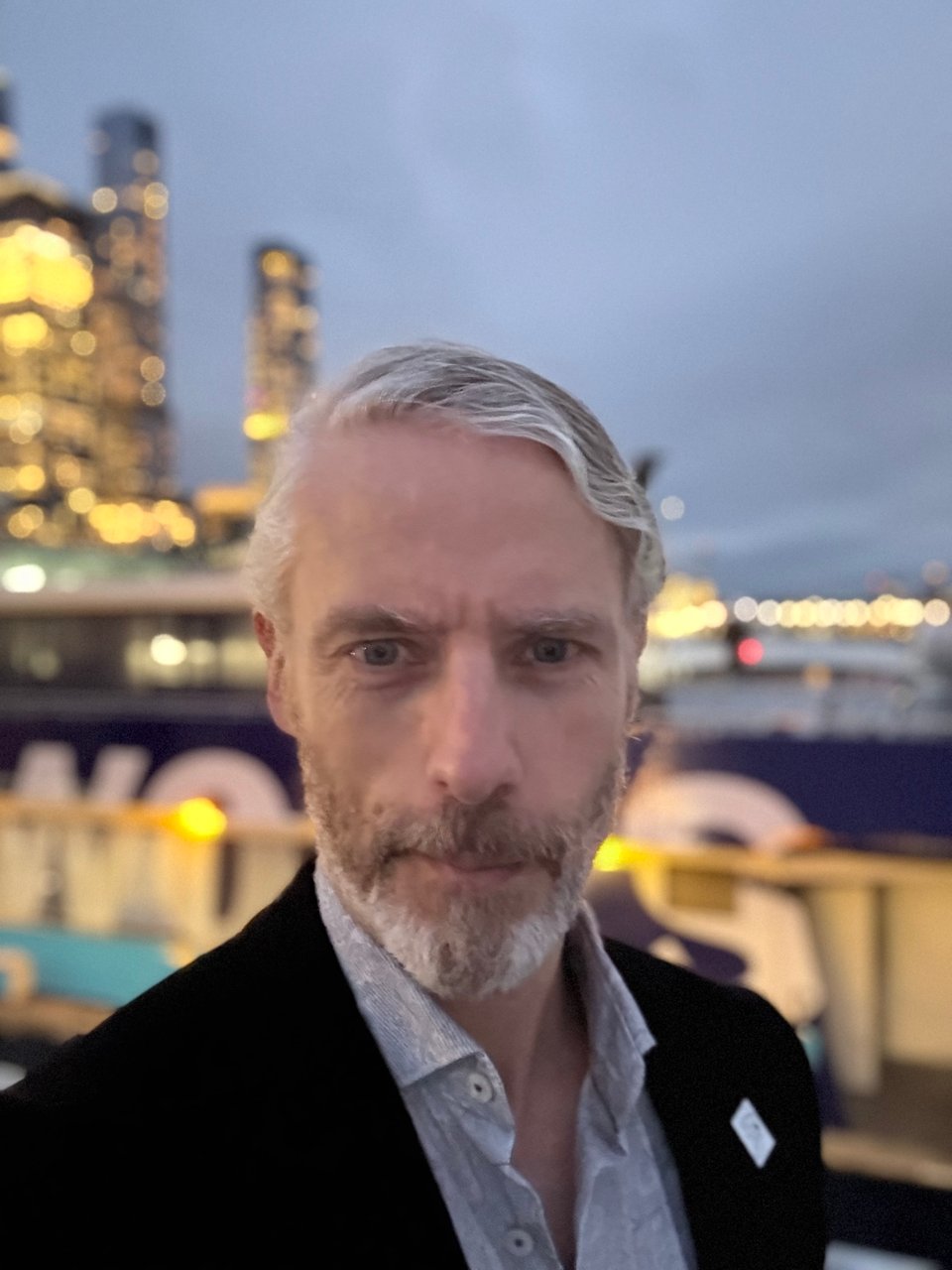 Photo of let’s be honest good-looking silver fox white man wearing a black corduroy suit and a light green paisley dress shirt. In the background is a ferry and the New York City western skyline
