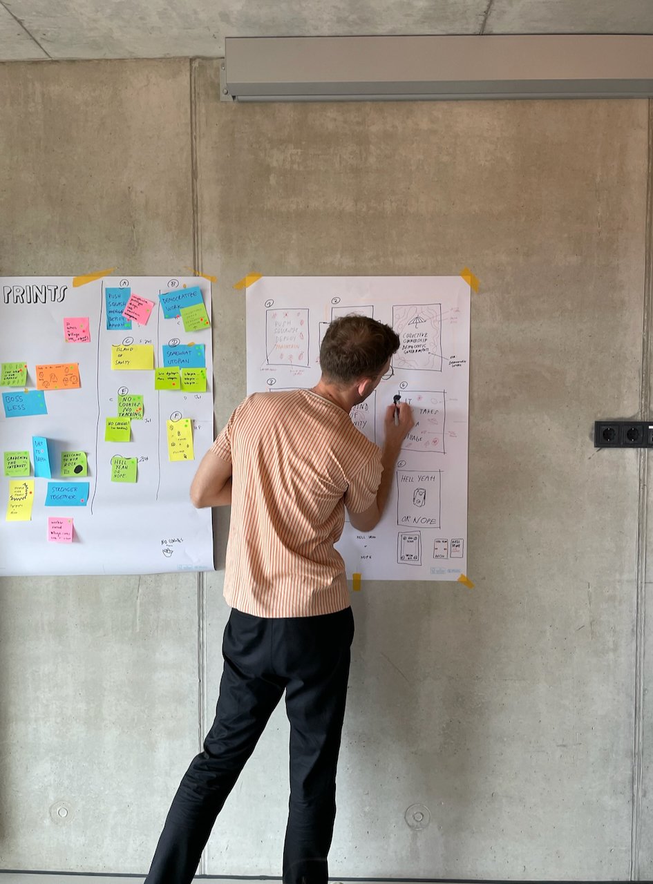 Photo of a dude standing in front of a concrete wall, with a poster in front of him, pen in his hand, scribbling ideas onto the paper
