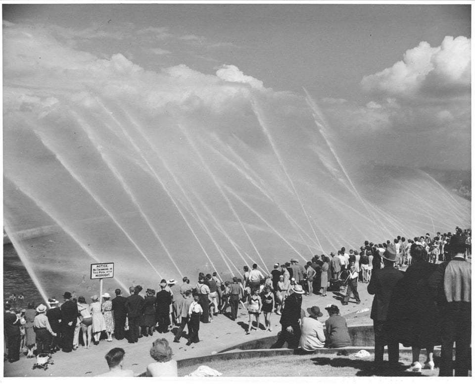 Black and white photo. A large group of people watch as many hoses spray water into the sky.