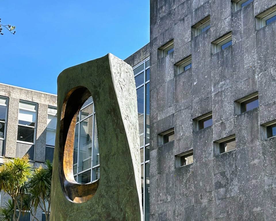 The rock form sculpture by Barbara Hepworth standing in the courtyard of Lys Kernow (county hall). The bronze sculpture has rough external sides that are turned green. Where it is pierced, the metal has been polished smooth and is bronze. The building beyond is concrete with fortress-like windows.