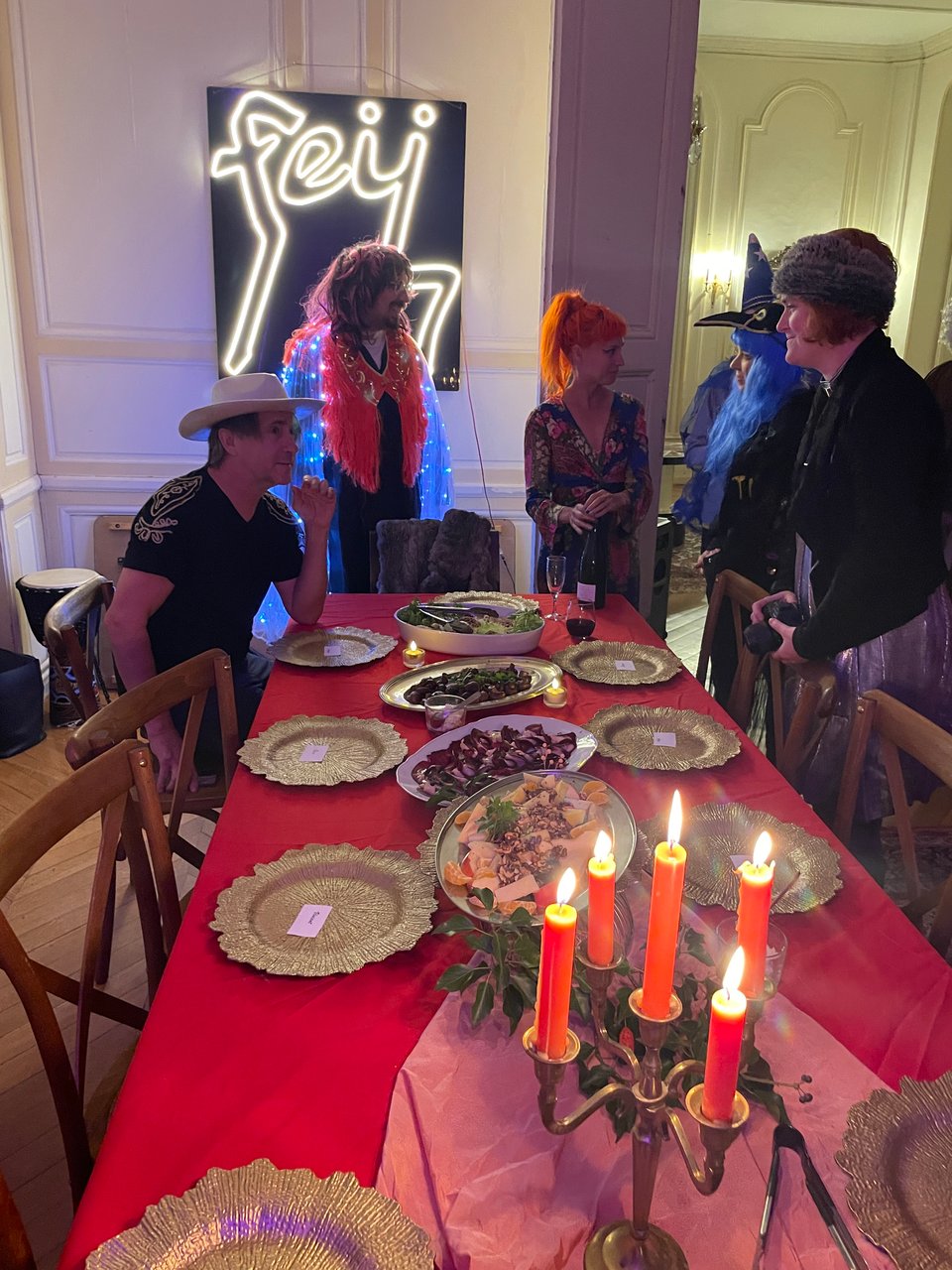 Five people are at the end of a long table which is covered in a red tablecloth, gold plates and there is a burning candelabra in the foreground.