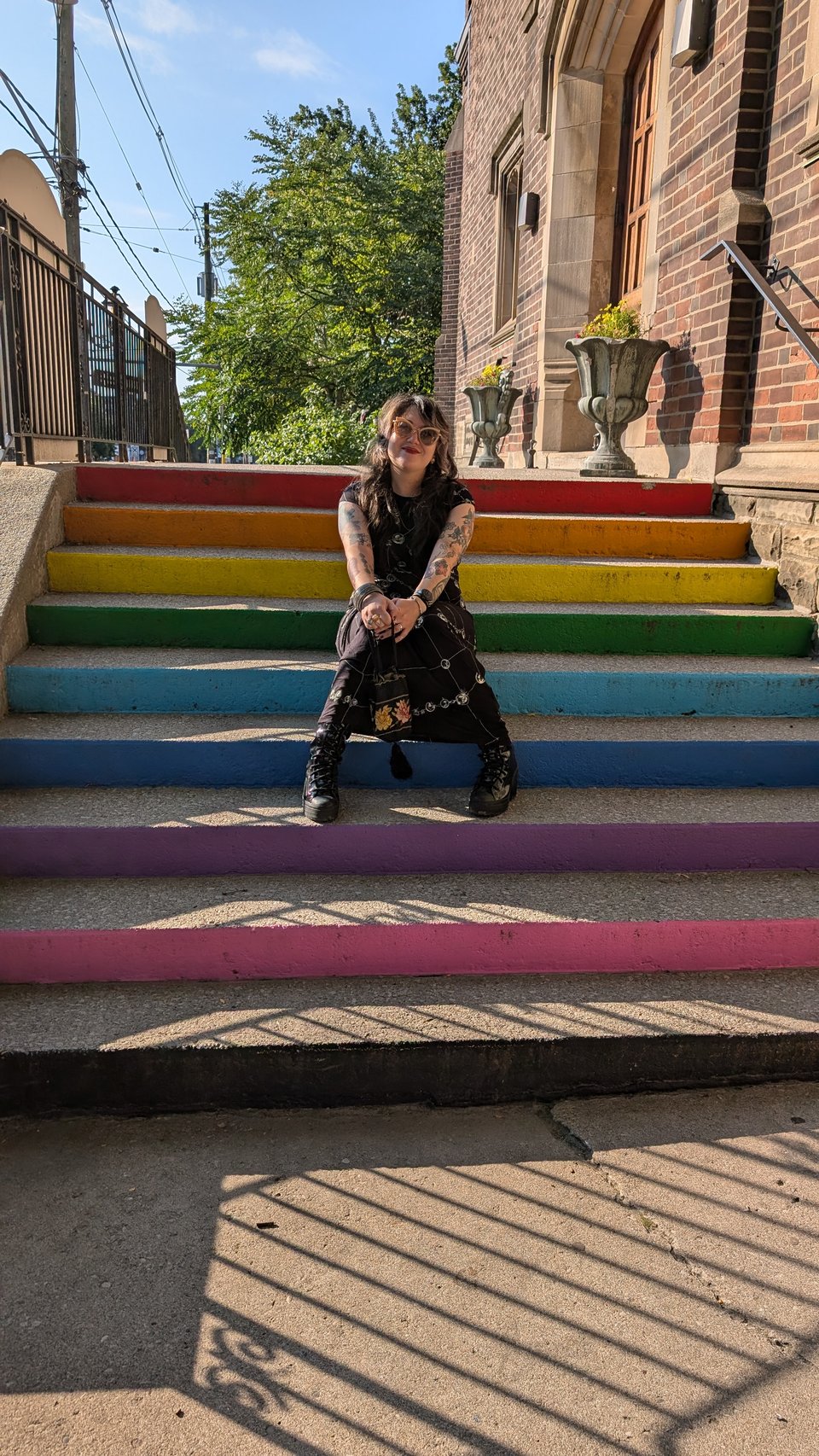 A white woman dressed in fashionable dark clothing and cute sunglasses poses, sitting on a set of concrete stairs painted in rainbow colors, one step for each color: red, orange, yellow, green, light blue, darker blue, lavender, and pink, for the gay pride flag.