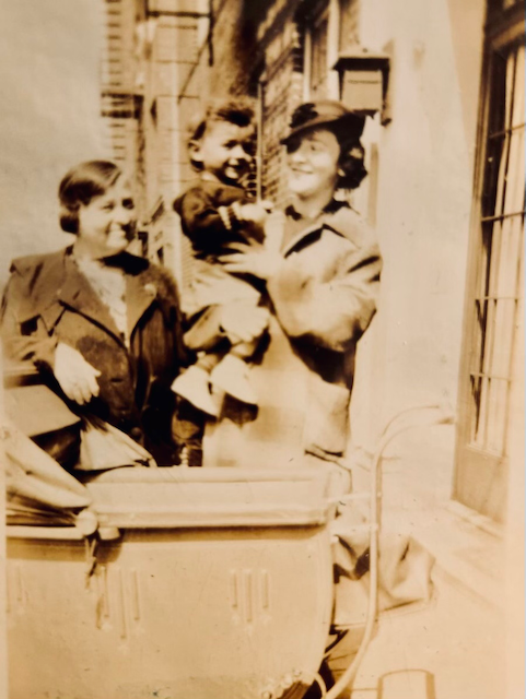Old black and white photograph, brown with age. Three people stand on the stoop of a New York tenement building. An older, shorter woman in a dark jacket, a taller, younger woman in a light coat, and a 2 or 3 year-old boy being held by the young woman. All are smiling, and the two women are looking at the child with love.