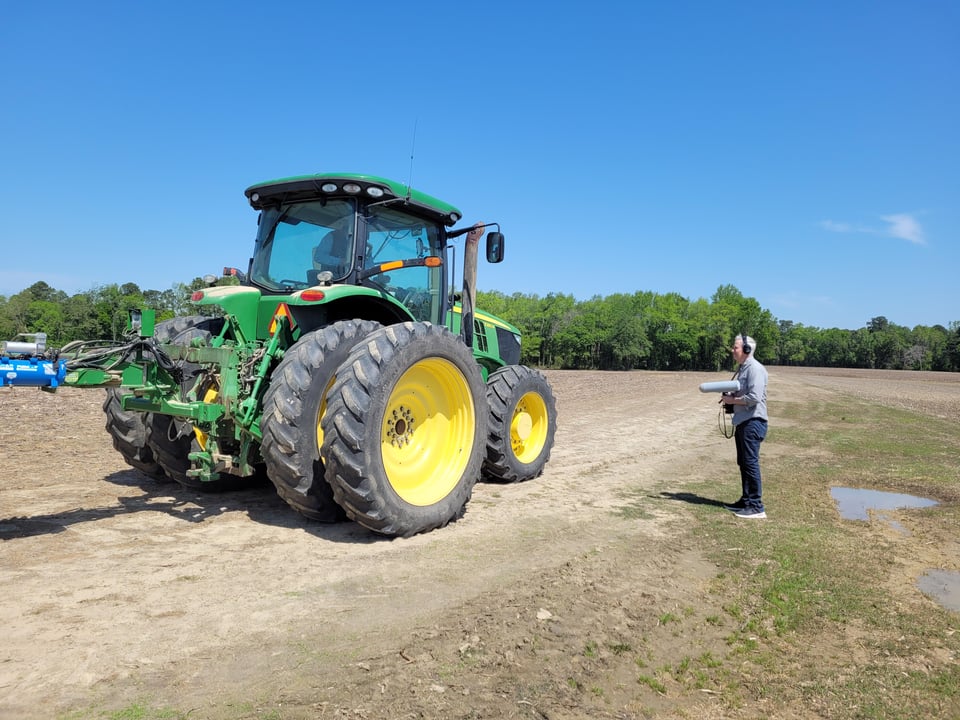 Photo of John Biewen standing in a field in headphones pointing his microphone at a large John Deere tractor.