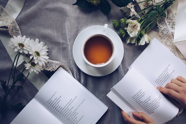Stock photo of a person whose hands are opening a book. There is also a cup of tea and some flowers on the table taht the book is resting on.