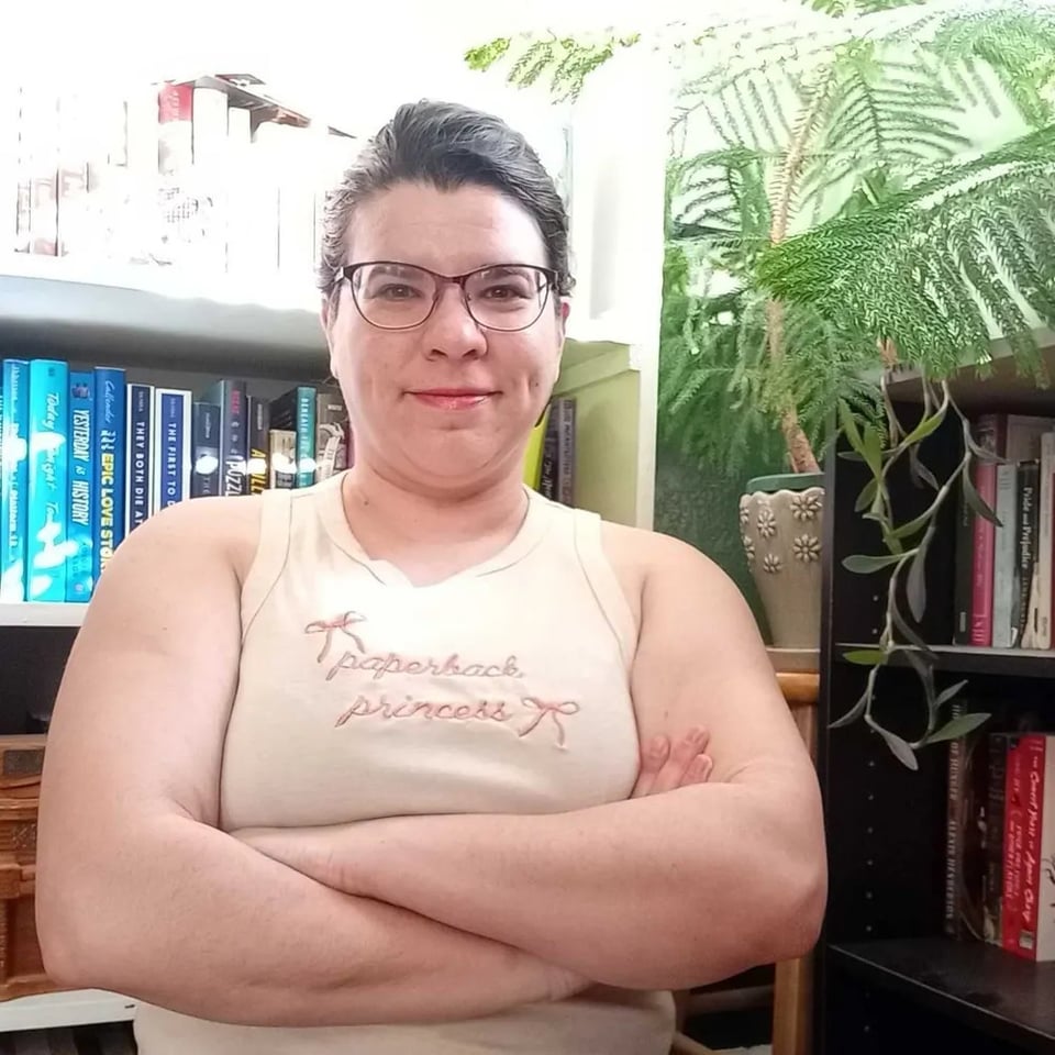 E sitting in front of two bookshelves with plants nearby, wearing a baby tank that says "paperback princess" and folding their arms, showing off frankly bulging arm muscles. buffity buff buff buff