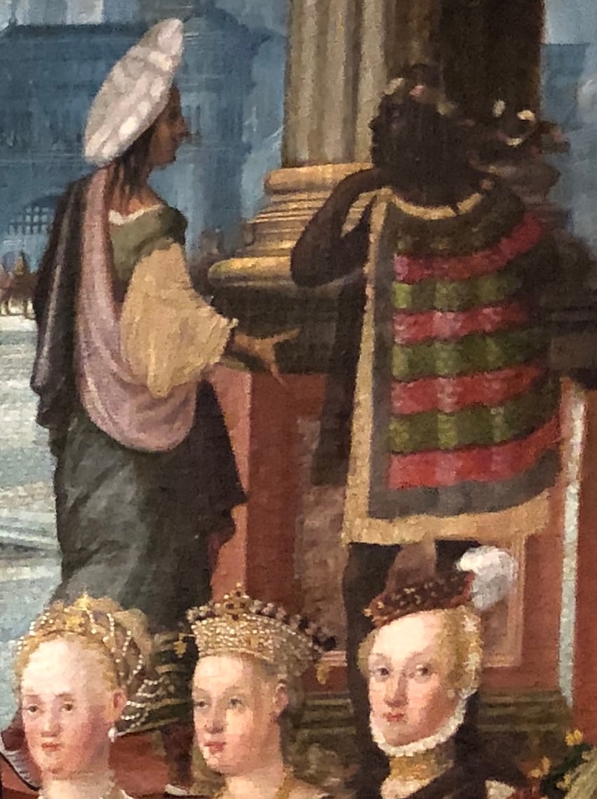 A dark-skinned figure in a green and crimson striped cape a gold border is in conversation with a lighter figure in robes and a hat. Along the bottom of the detail are three heads of white women in fine outfits (just visible at the neck) and hats/tiaras.