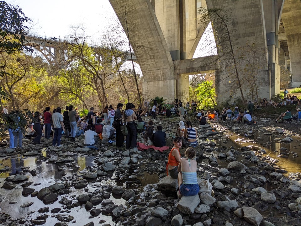 A crowd of people gather under the arches of the Colorado Street Bridge in Arroyo Seco, watching a synthesizer concert on the opposite bank. The river is low and muddy, with rocks poking up out of the water. People sit on rocks or on blankets in the mud. Beyond the bridge, the light green trees are lit by afternoon sunlight.