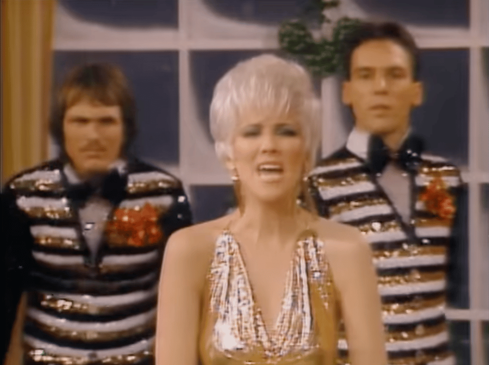 Catherine O'Hara with a short blond pussycat wig and silver lamé dress as sexy lounge singer starlet Lola Heatherton, flanked by two of her faithful backup dancers.
