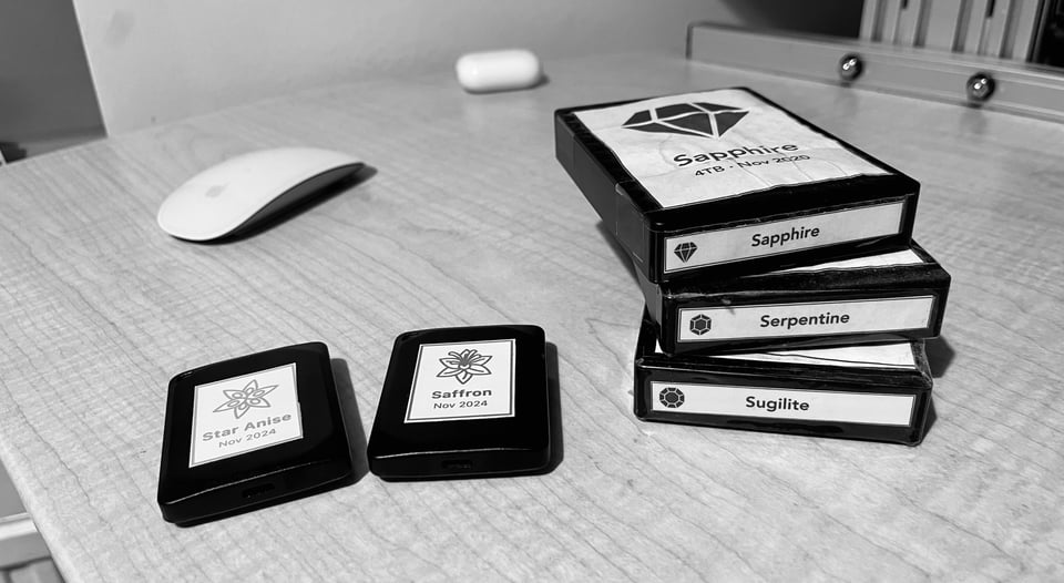 Black and white photo of two piles of disk. On the right-hand side are large black boxes labelled “Sapphire”, “Serpentine” and “Sugilite”. On the left-hand side are two significantly smaller black boxes, labelled “Star Anise” and “Saffron”.