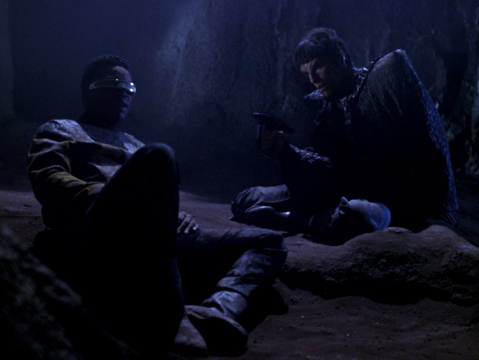 Geordi and a Romulan officer stuck in a muddy pit. The Romulan has a disrupter weapon pointed towards Geordi, who is seated and resting.