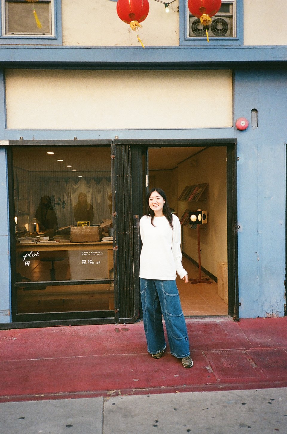 Alice in front of the open door and large storefront window. The vinyl on the window reads Plot in hand-drawn cursive with the Chinese symbol below it. Inside you can see warm light, display shelves, a large table filled with objects and a curtain in the back.