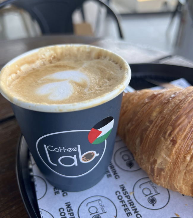 A paper coffee cup with the logo of Coffee Lab and a round decal of a Palestinian flag, on a plate with a croissant