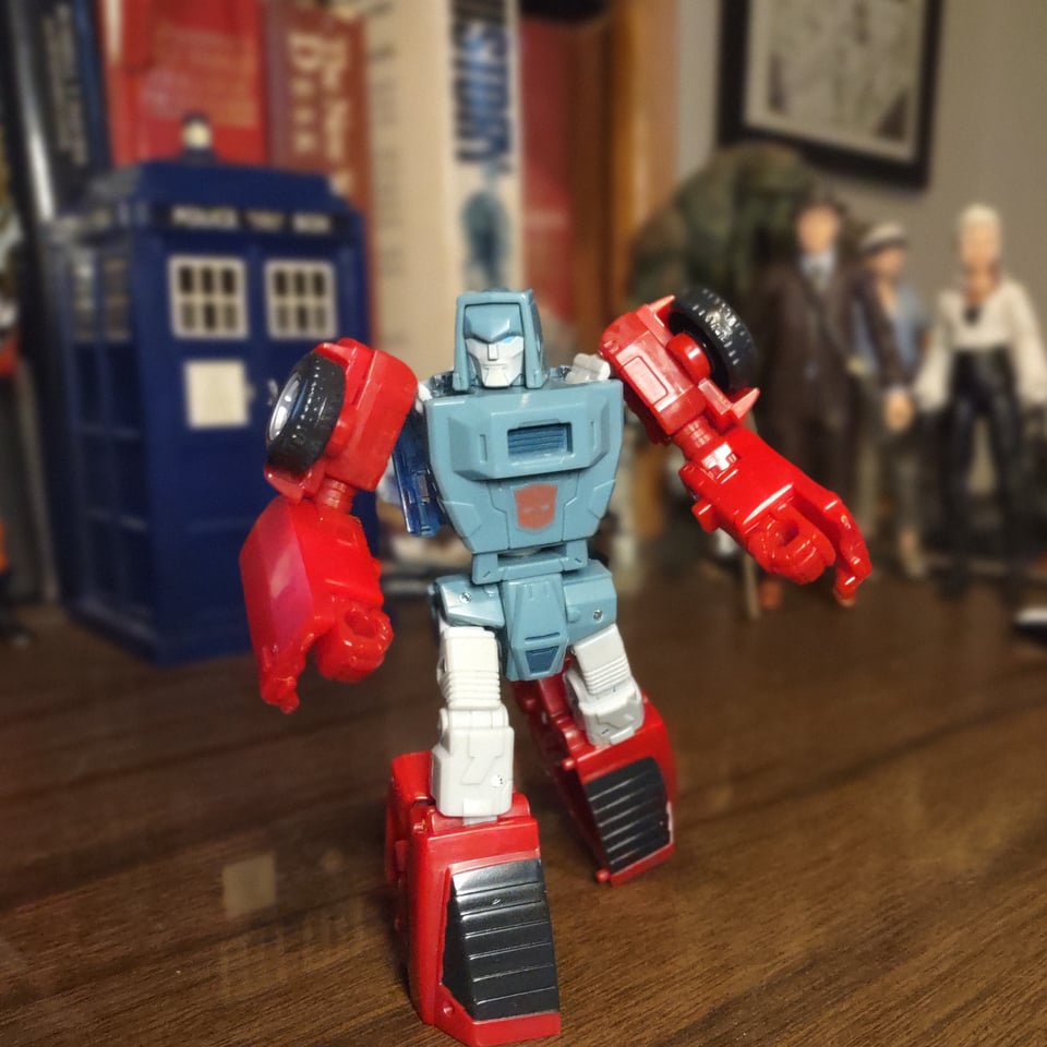 A red and gray Transformer stands on a wooden desk, with other action figures in the background. He has a squared-off head and wheels on his shoulders. A red Autobot  crest is centered on his torso.