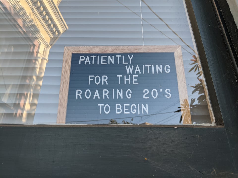 A sign in someone's window that reads "PATIENTLY WAITING FOR THE ROARING 20s TO BEGIN"