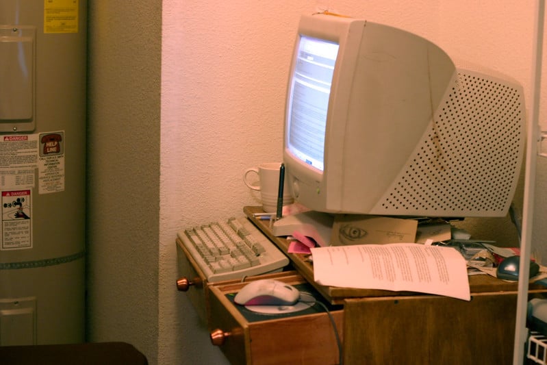 A photo of a CRT screen monitor on a janky wooden desk, drawers pulled out and upside down to create a space for a mouse and a keyboard. Papers are strewn around the monitor, a water heater is visible in a nook to the left of the photo, by where a person would sit.