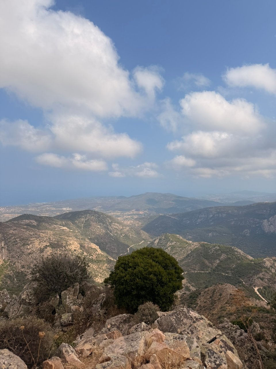 A panoramic view across rugged, rocky mountains and green valleys beneath a blue sky filled with large white clouds.