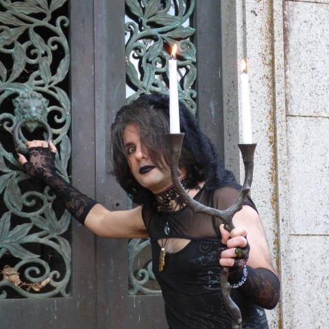 Bio picture of Bitter Karella, she's holding a candlelabra in front of a gothic looking gate wearing gothic clothing