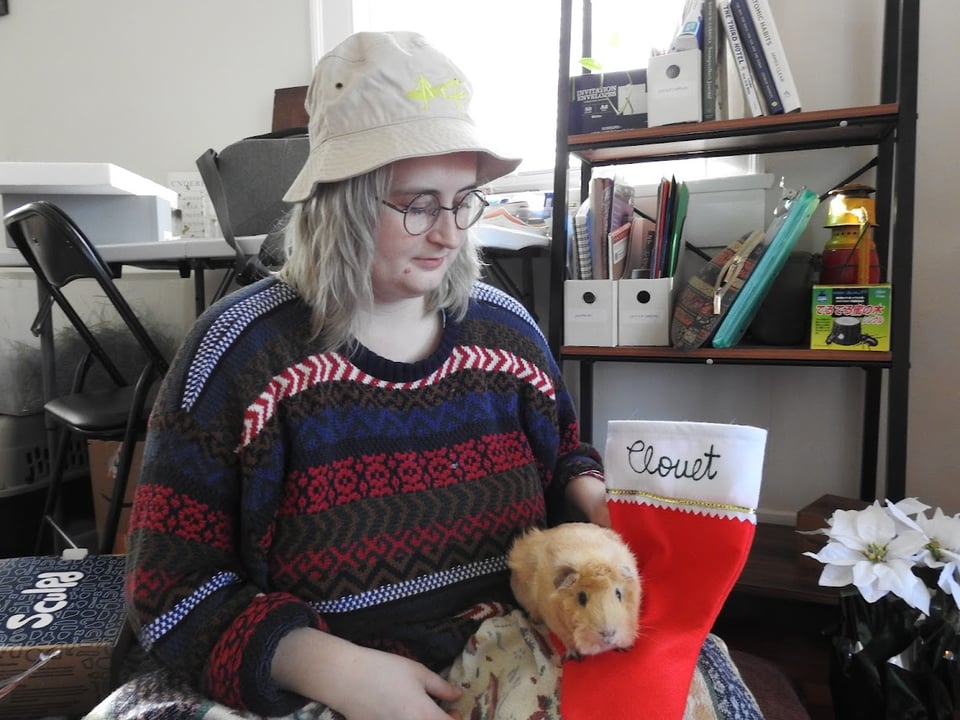 Maria, a white person with platinum blonde hair, wears an ugly Christmas sweater and a tan bucket hat with a mantis on it. They hold Clouet the guinea pig as she shows off her Christmas stocking, which has "Clouet" spelled out in green puff paint.