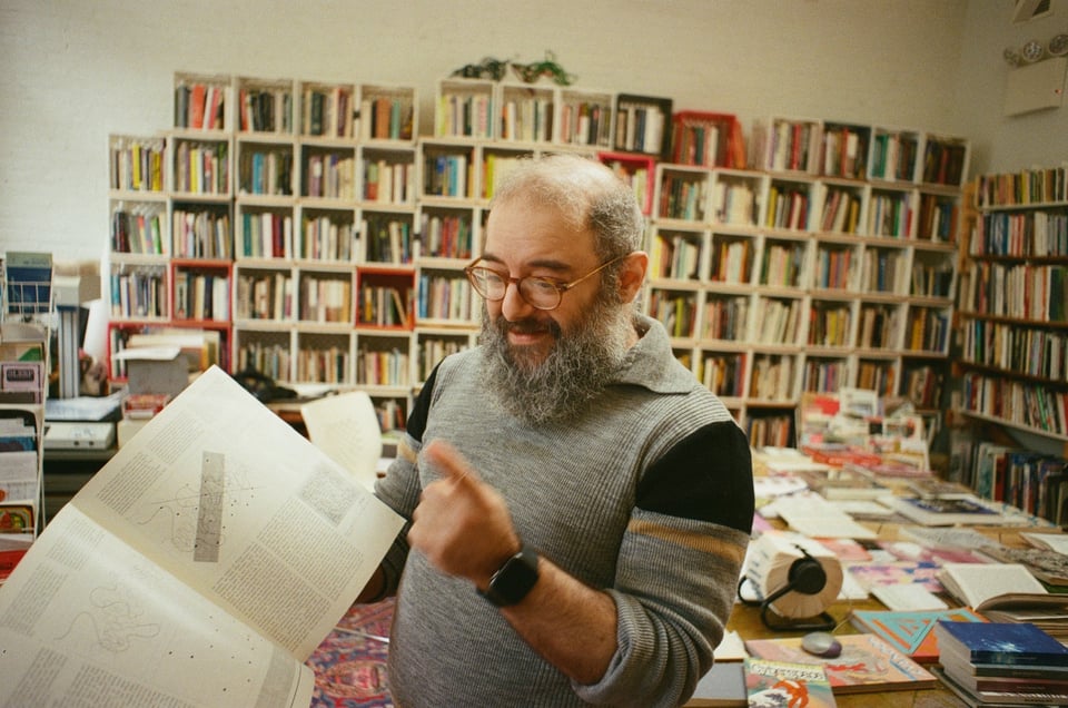 [David holding an open book and pointing at something within its pages. The library shelves, a table covered in books, and various machines like a scanner can be seen in the background.]