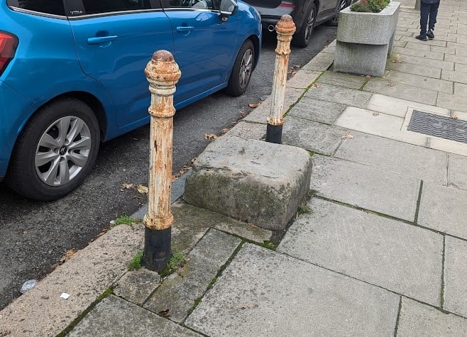 A square stone a bit more than a foot across, sticking out about half a foot from a pavement, with some rusty bollards either side.