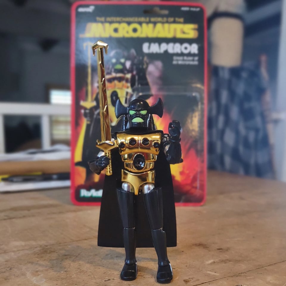 The Emperor action figure stands on a wooden table. He has a black helmet (with large green eyes), arms and legs, with a gold torso and sword. Also a black vinyl cape. The cardback he came on is behind him--with its big retro MICRONAUTS logo, and the action figure's in roughly the same pose as the card art.