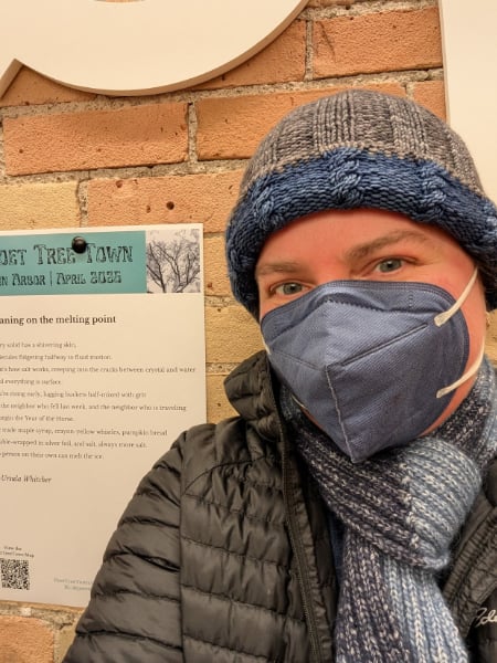 A person wearing a blue and gray cable-knitted hat, a blue mask, and a blue striped scarf stands in front of a broadsheet for PoetTreeTown. Part of the Spun logo is visible in the background.