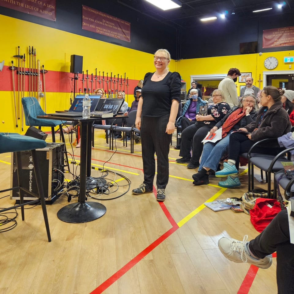 Kate standing in a hall with swords on the wall and a radio desk set up, and the audience in their seats behind her. Her expression is WHAT IS MY LIFE.