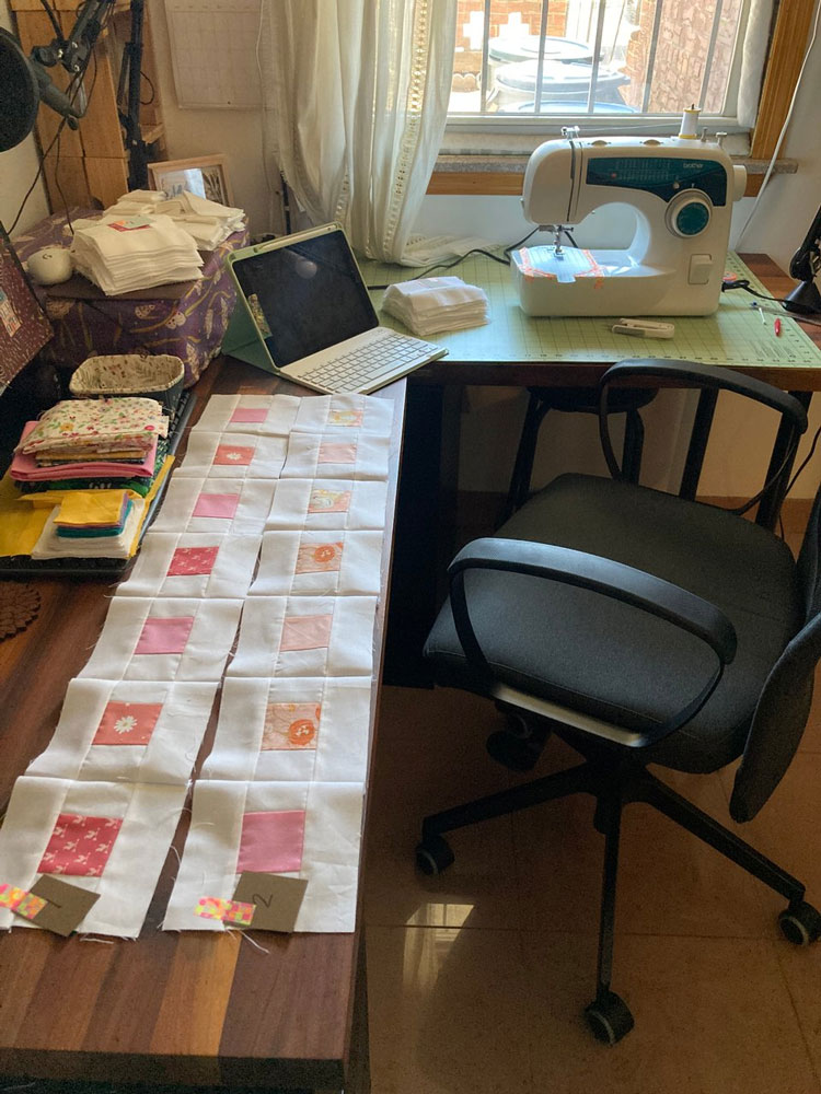 Two rows of quilt blocks sewn together, showing a range from red to peach scraps encompassed in white squares. The rows rest on a wooden desk which has all kinds of other things on top of it: a tablet, a few stacks of scrap fabric for a different project, a large green cutting mat under a sewing machine, a small basket to catch threads, more quilt blocks waiting to be sewn, a computer, and a printer. Next to the desk is a window.