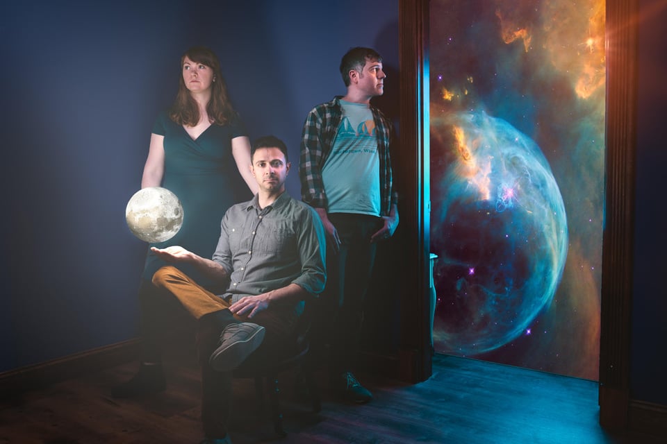 The three members of True Jackie pose, with one of them holding his hand underneath a small, levitating moon, while another looks at the Earth spinning in the cosmos.