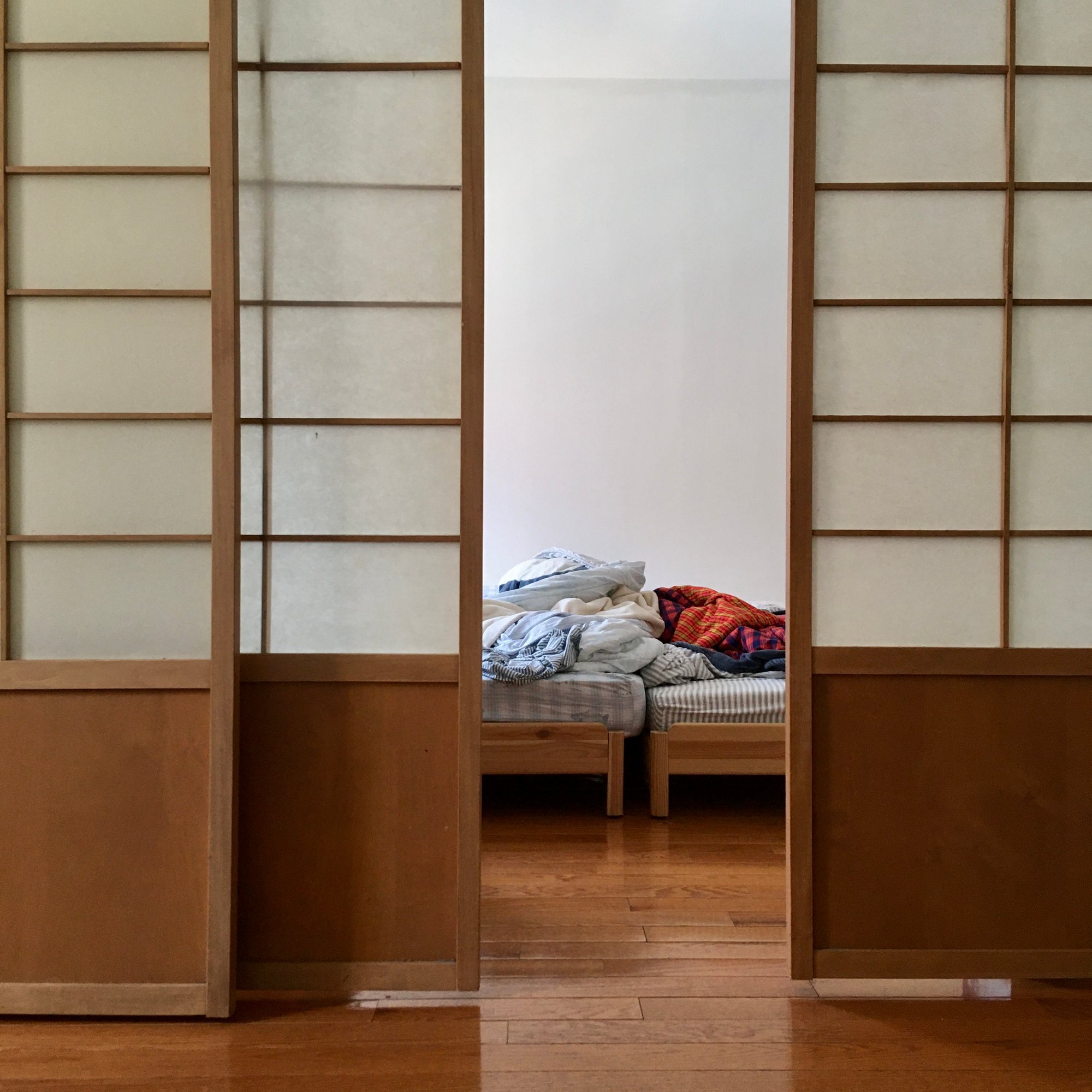 Photo of two low beds, half-obscured by three shoji panels. A mess of clothes and sheets lie atop the beds.