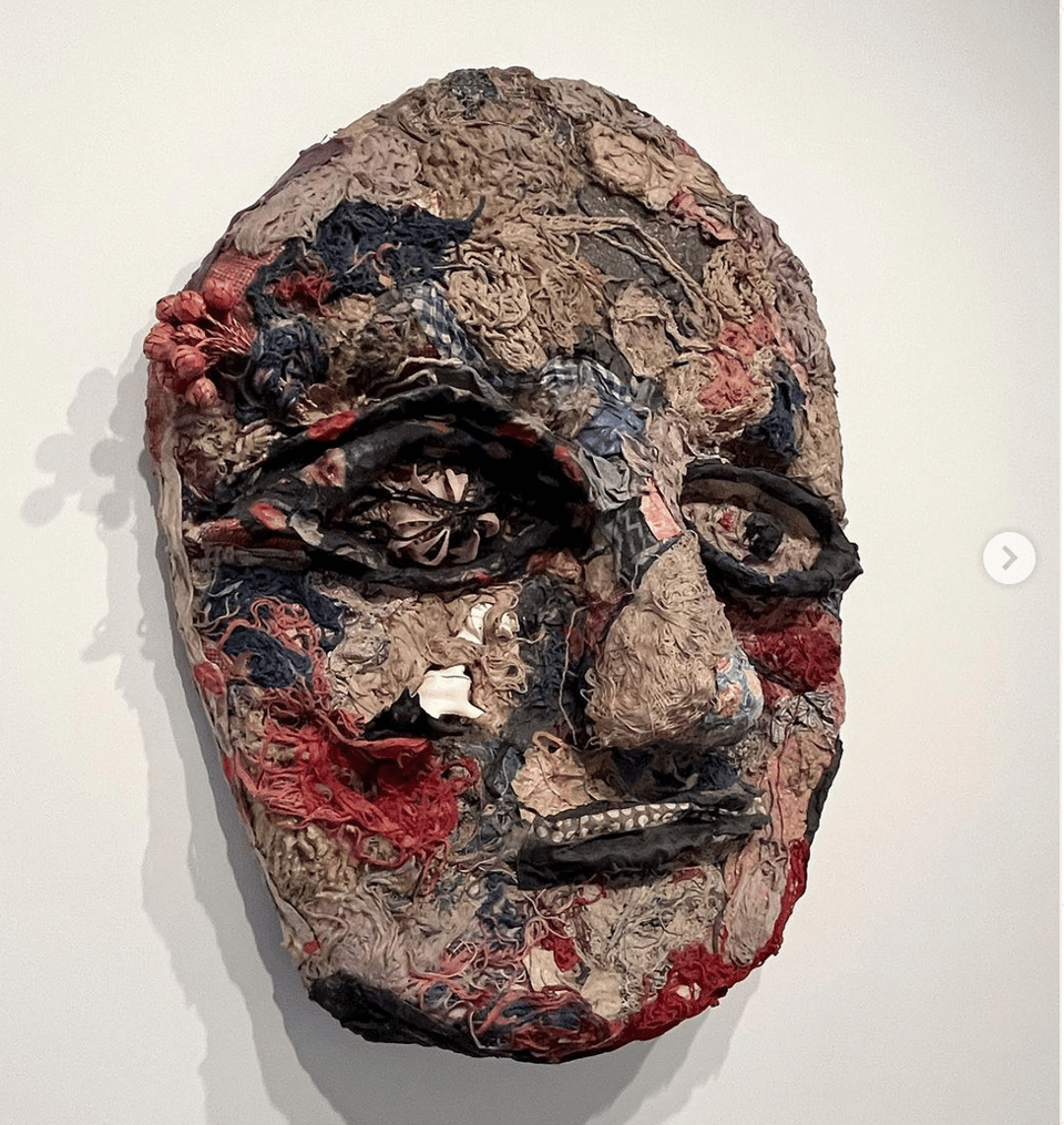 an unusual mask made from tiny scraps of different old fabrics and threads, mounted on a gallery wall