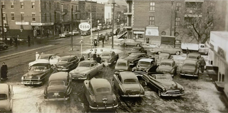 Black and white 1950's photo of an Esso gas station with many cars parked on its lot.