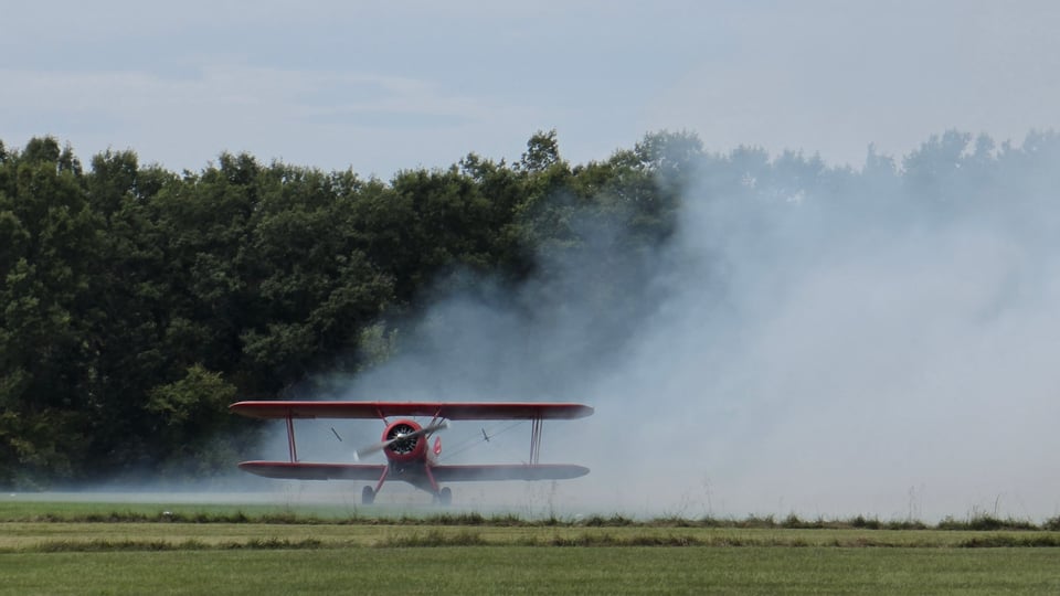 A red biplane sits on a grass airfield, its propeller spinning as thick white smoke billows across the field and drifts up in front of a dense line of dark green trees under a pale sky.
